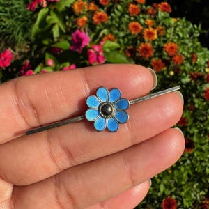 Könnte beinhalten: Eine silberfarbene Stab-Brosche mit einem zentralen Blumenmotiv, das aus acht leuchtend blauen Emaille-Blütenblättern und einer kleinen, dunklen, texturierten kreisförmigen Mitte besteht. Der Metallstab verläuft horizontal.