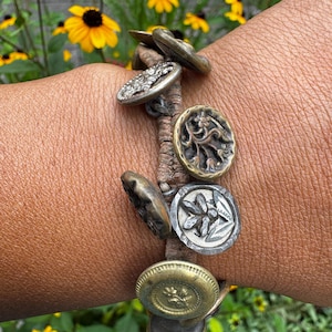 May include: A bracelet featuring a series of assorted metallic buttons in various shades of brass, silver, and bronze, each displaying unique embossed floral and geometric patterns, securely attached to a textured, light brown woven cord band.