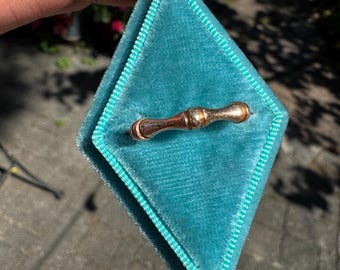 May include: A small, antique gold-toned ring with a cylindrical design, resting inside a teal velvet ring box. The ring box is diamond-shaped with a teal border. The background is a blurred outdoor scene.
