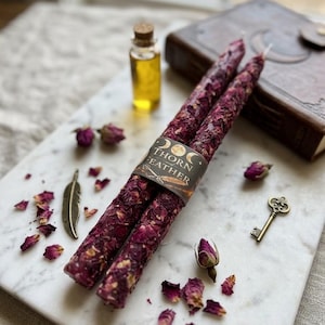 May include: Two long taper candles covered in deep red dried rose petals, bound by a paper label reading Thorn and Feather with moon icons. Nearby are a small glass vial of golden oil, a metal feather charm, a small key, and loose rose buds.