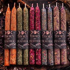 May include: A collection of colorful, decorative spell candles. The candles are wrapped with a black label that reads "THORN and FEATHER". The candles are various colors, including orange, green, red, and purple. A lit candle is in the background.