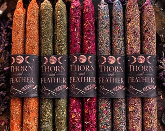 May include: Ten taper candles in five distinct color pairs, including orange, green, magenta, blue, and purple. Each candle is coated in dried herbs and petals. A black paper band wraps around each pair, featuring the text Thorn and Feather.