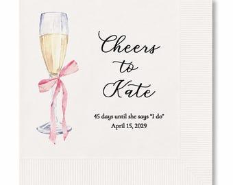May include: A square white paper napkin featuring a watercolor illustration of a champagne flute tied with a pink ribbon. The text reads: Cheers to Kate, 45 days until she says I do, April 15, 2029. The napkin has a textured border.