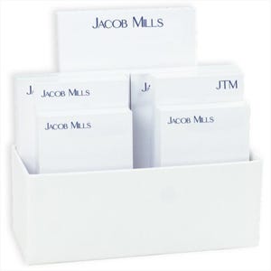May include: A white display case filled with white notepads. The notepads are stacked in a tiered arrangement, with the text "JACOB MILLS" printed in navy blue on each notepad. The display case is also white.