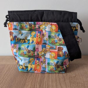 May include: A drawstring bag with a colourful patchwork design featuring characters from The Simpsons cartoon series. The bag has a black top and strap, and is on a wooden surface.