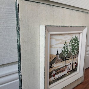 May include: A framed artwork featuring a village scene with figures, trees, and a church. The artwork is set within a white frame with a distressed, off-white outer frame. The artwork is signed in the lower right corner.