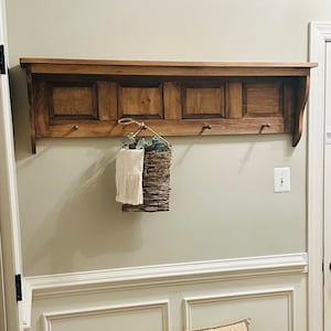 May include: A wooden coat rack with a shelf and hooks. The rack is stained a dark brown color and has a decorative design on the front. A small basket with a white towel is hanging from one of the hooks. A bench with a horse-themed pillow is in front of the rack.
