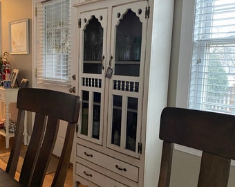 May include: A white wooden cabinet with glass doors and drawers. The cabinet has a distressed finish and is decorated with ornate details. The cabinet is in a room with a wooden floor and white walls.
