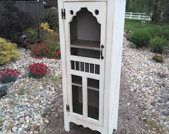 May include: White distressed wood cabinet with a glass door and a shelf inside. The cabinet has a decorative arched top and a scalloped base.