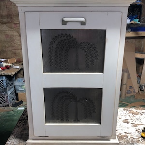 May include: White painted wooden cabinet with two doors. Each door has a metal panel with a cutout design of a weeping willow tree. The cabinet has a simple handle.