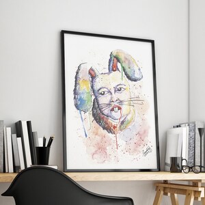 Puede incluir: Una pintura de acuarela enmarcada de una figura parecida a un conejo con acentos coloridos. La obra de arte presenta un marco negro y se exhibe en un escritorio de madera. El escritorio también contiene libros, una silla negra y una ventana con vistas.