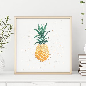 Könnte beinhalten: Aquarellmalerei einer Ananas in einem goldenen Rahmen. Die Ananas ist gelb und orange mit grünen Blättern. Das Kunstwerk wird auf einer weißen Oberfläche mit einer Vase mit Grün und einem Stapel Bücher ausgestellt.