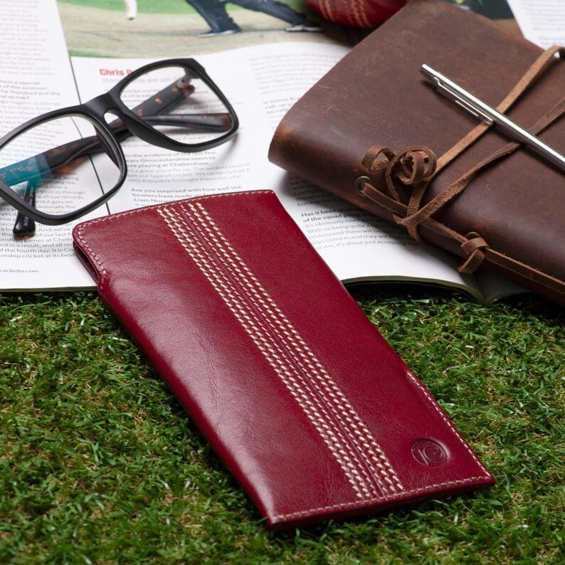 May include: A burgundy leather wallet with white stitching in the shape of cricket seams. The wallet has a circular logo with the letter "C" inside.