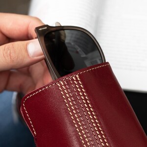 May include: A burgundy leather sunglasses case with white stitching. The case has a stitched design that resembles a cricket ball.