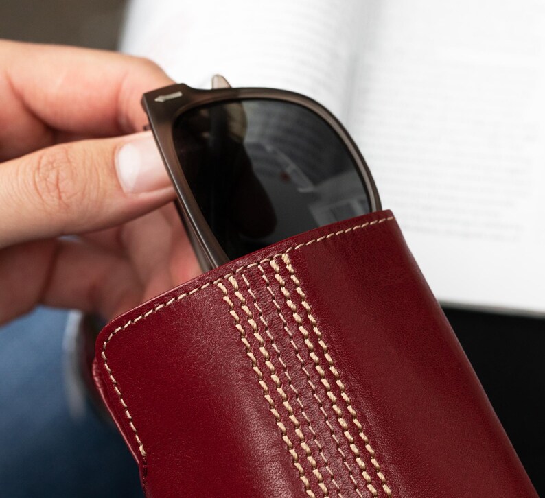 May include: A burgundy leather sunglasses case with white stitching. The case has a stitched design that resembles a cricket ball.