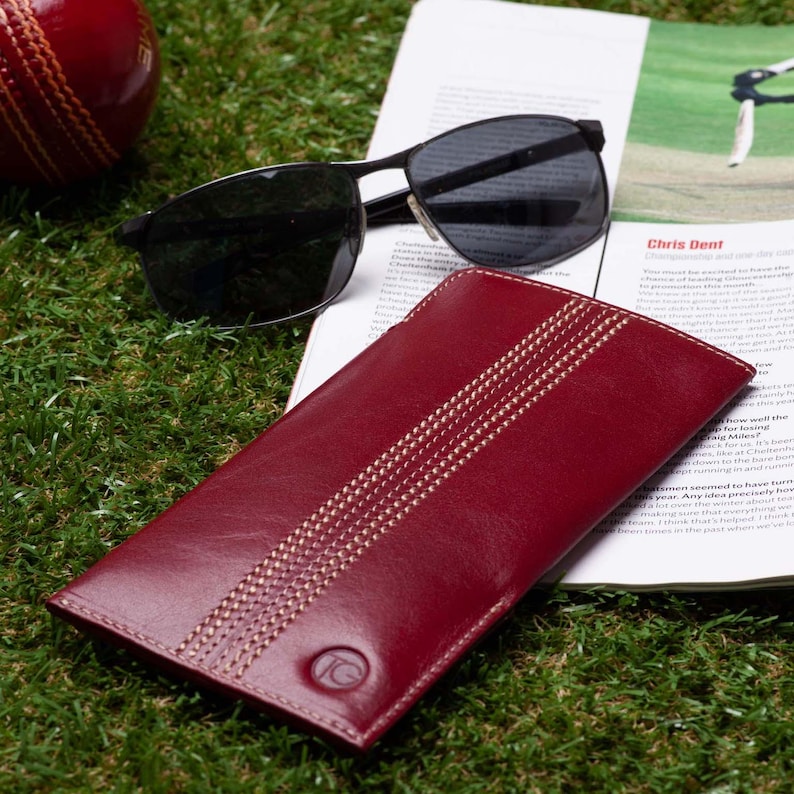 May include: A red leather sunglasses case with white stitching that resembles a cricket pitch. The case has a small gold circle with the letter "C" in the center.