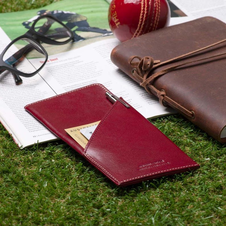 May include: A red leather passport holder with a pen holder and a card slot. The holder is lying on a green grassy surface with a magazine and a brown leather journal in the background.
