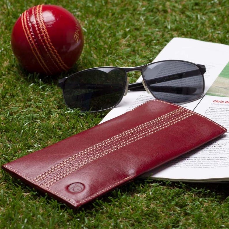 May include: A red leather glasses case with white stitching and a cricket ball design. The case is lying on a magazine with sunglasses on top.