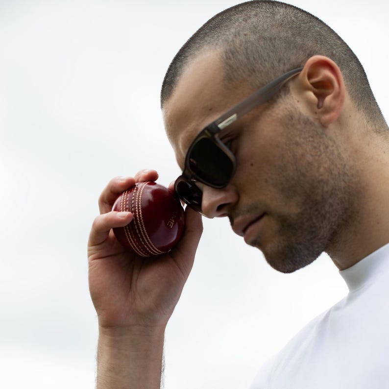 May include: A man wearing brown sunglasses holds a red cricket ball in his hand. He is looking down at the ball.