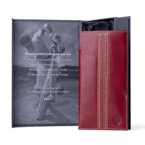 May include: A red leather wallet with white stitching, featuring a cricket pitch design. The wallet is in a box with a cricket player on the front. The text on the box reads "Because cricket is the game you love. The distinctive sound of leather on willow, the smell of freshly cut grass, the feel of the ball in your hands, it's the passion we have for cricket. Visit us at www.thegame.cool Proudly supporting the McGrath Foundation."