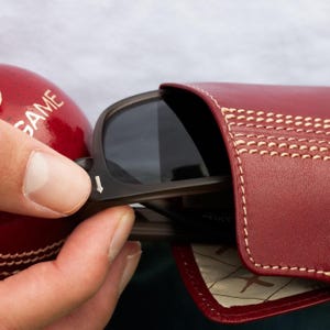 May include: A red leather sunglasses case with white stitching. The case has a cricket ball design and is open to reveal a pair of sunglasses.