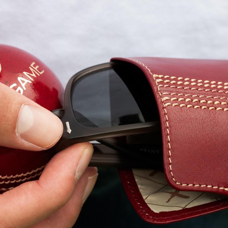 May include: A red leather sunglasses case with white stitching. The case has a cricket ball design and is open to reveal a pair of sunglasses.