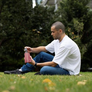 May include: A person sitting on the grass, wearing blue jeans and a white t-shirt, holding a burgundy sunglasses case.