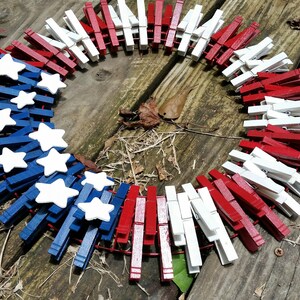 以下が含まれることがあります： 洗濯バサミで作った赤、白、青の愛国的なリース。リースは白い星で飾られています。