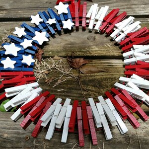 以下が含まれることがあります： 木製の洗濯ばさみで作った赤、白、青のアメリカ国旗のリース。洗濯ばさみはアメリカ国旗に似せて塗られています。