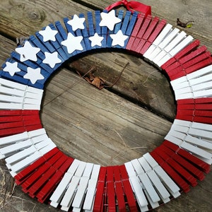 May include: A red, white, and blue American flag wreath made from wooden clothespins. The wreath is decorated with white stars.