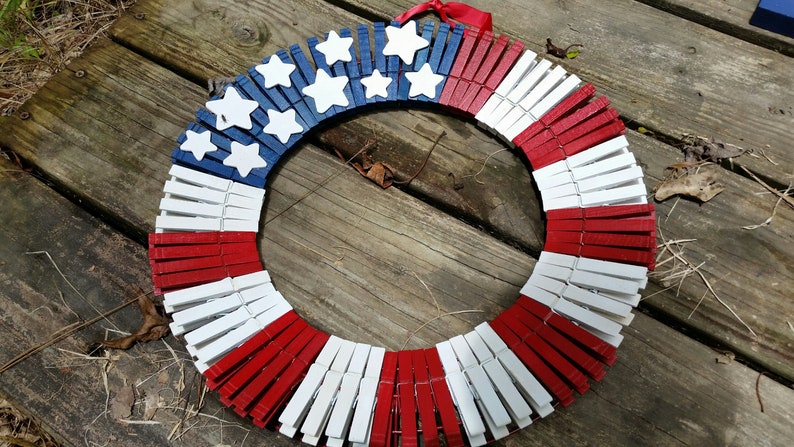 May include: A red, white, and blue American flag wreath made from wooden clothespins. The wreath is decorated with white stars.