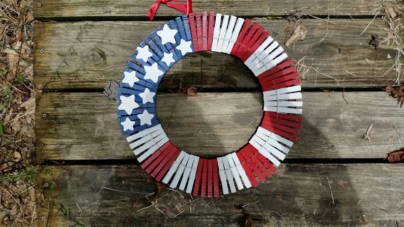 May include: A red, white, and blue American flag wreath made from clothespins. The wreath is decorated with white stars on a blue background.