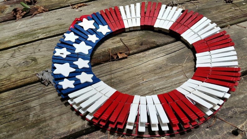 May include: A red, white, and blue patriotic wreath made from wooden clothespins. The wreath is decorated with white stars on a blue background.