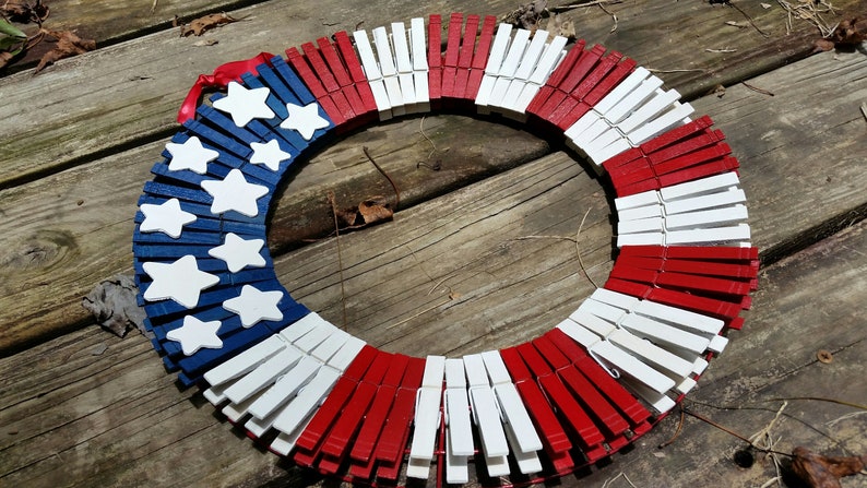 May include: A red, white, and blue patriotic wreath made from wooden clothespins. The wreath is decorated with white stars on a blue background.