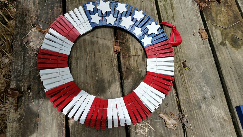 May include: A red, white, and blue wreath made from clothespins. The clothespins are painted to resemble the American flag. The wreath is decorated with white stars.