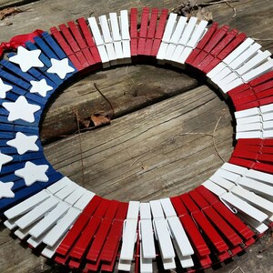 May include: A red, white, and blue American flag wreath made from wooden clothespins. The clothespins are painted to resemble the American flag.
