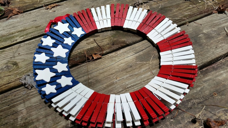 May include: A red, white, and blue American flag wreath made from wooden clothespins. The clothespins are painted to resemble the American flag.