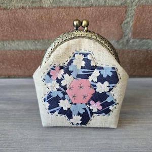Op de afbeelding: Een beige portemonnee met een bronskleurige metalen knipsluiting. De voorzijde heeft een centrale roze zeshoek omringd door zes donkerblauwe zeshoeken met een lichtblauw en wit bloemmotief, allemaal met zichtbaar garen op de stof gestikt.