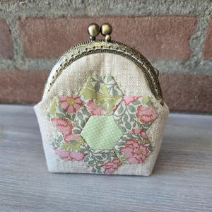 Pode incluir: Uma pequena bolsa para moedas feita de tecido de linho bege com um design central de patchwork de hexágonos florais em tons de rosa, verde e branco. A bolsa possui uma estrutura de metal cor de latão antigo com fecho de clique.