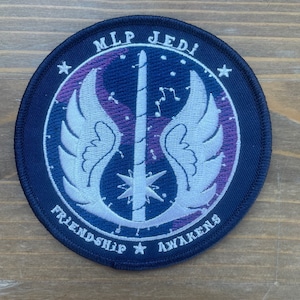 Puede incluir: Un parche redondo bordado azul con un símbolo blanco de sable de luz y alas. El texto "MLP Jedi" está en la parte superior del parche y "Friendship Awakens" está en la parte inferior.