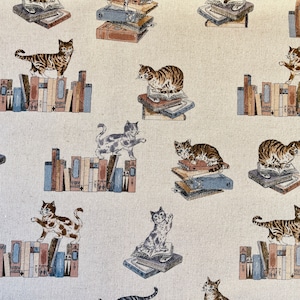 Puede incluir: Una tela beige con un patrón repetido de gatos sentados en pilas de libros. Los gatos son marrones, negros y blancos con marcas de atigrado. Los libros son marrones, azules y rojos.
