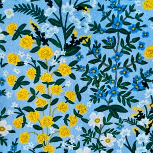 May include: A blue fabric with a floral pattern featuring yellow roses, blue forget-me-nots, and white flowers. The fabric has a subtle linen texture. "CAMONT" and "RIFLE PAPER CO." are printed on the left side of the fabric.
