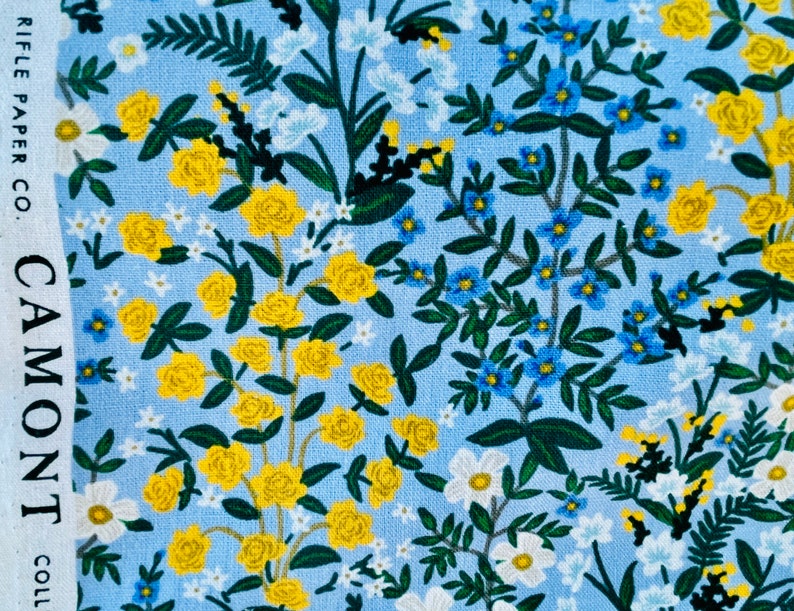 May include: A blue fabric with a floral pattern featuring yellow roses, blue forget-me-nots, and white flowers. The fabric has a subtle linen texture. "CAMONT" and "RIFLE PAPER CO." are printed on the left side of the fabric.