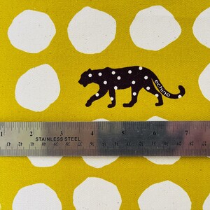May include: Yellow fabric with white polka dots and a black silhouette of a leopard with white spots. The leopard is walking to the right and has the word "echino" printed on its side. A ruler is placed below the fabric.