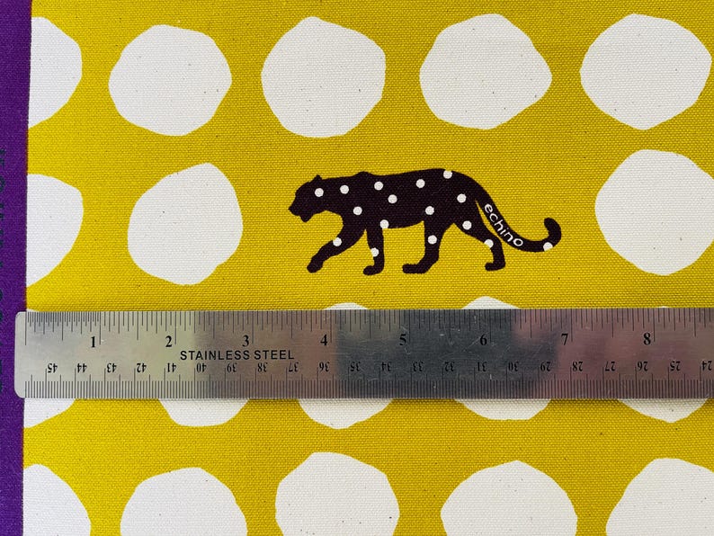 May include: Yellow fabric with white polka dots and a black silhouette of a leopard with white spots. The leopard is walking to the right and has the word "echino" printed on its side. A ruler is placed below the fabric.