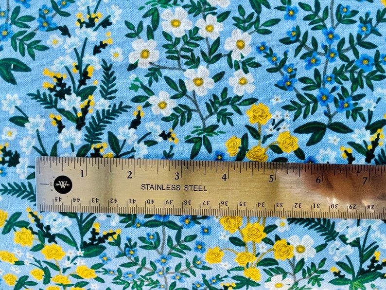 May include: A blue fabric with a white and yellow floral pattern. The fabric is measured with a ruler that reads "STAINLESS STEEL".