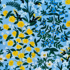 May include: A blue fabric with a floral pattern featuring yellow roses, blue forget-me-nots, and white flowers. The fabric has a subtle linen texture. "CAMONT" and "RIFLE PAPER CO." are printed on the left side of the fabric.