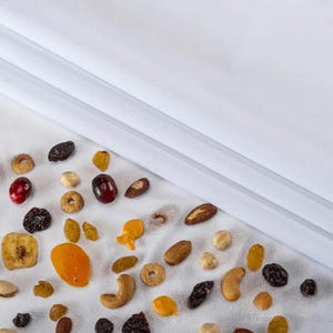 May include: A white fabric surface with a variety of dried fruits and nuts scattered on it. The nuts include almonds, cashews, and peanuts. The dried fruits include apricots, cranberries, and raisins.