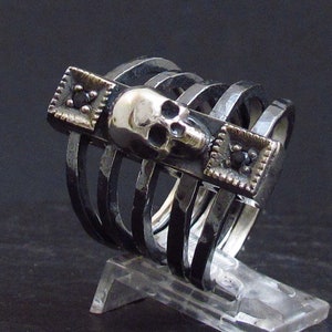 Puede incluir: Un anillo de plata con un diseño de calavera y piedras preciosas negras. El anillo está hecho de múltiples bandas que se apilan una encima de la otra.
