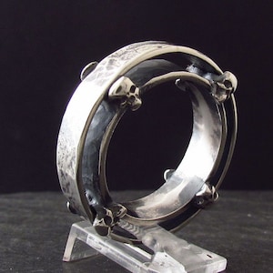 May include: A silver ring with a textured band and skull-shaped accents. The ring is made of two bands that are joined together.